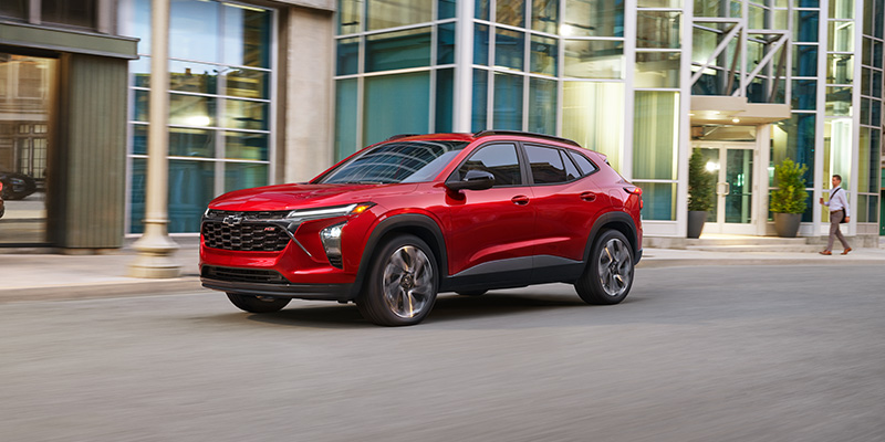 Front-three-quarter view of a bright red Chevrolet Trax driving down a city street in front of modern glass buildings.
