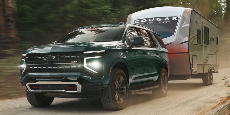 A dark green Chevrolet Tahoe Z71 towing a large Cougar travel trailer along a dirt road through a forest.